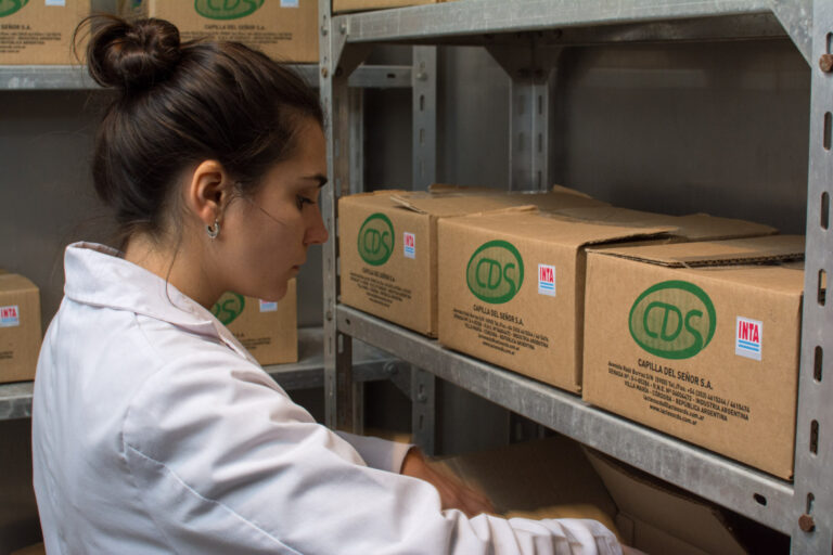 Una persona con guardapolvo junto a estantes con cajas identificadas con las marcas CDS e INTA.